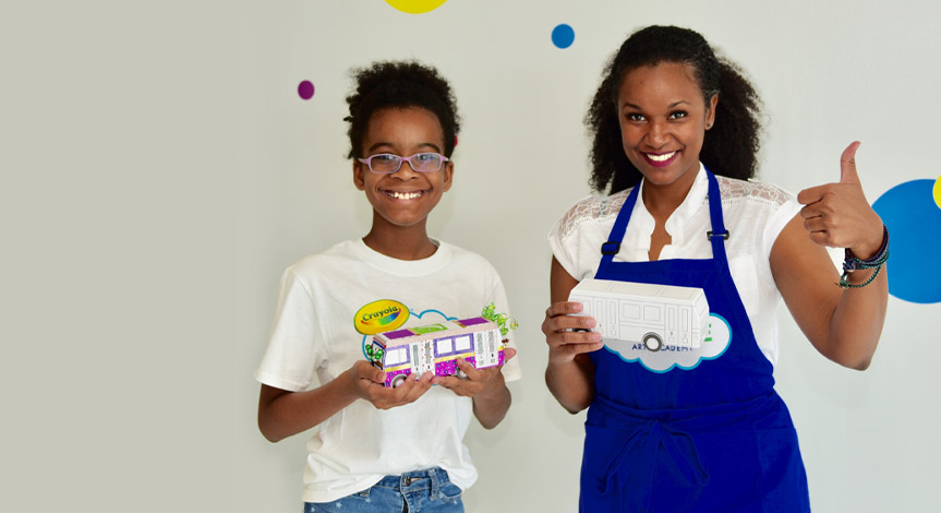 A girl holding her bus she made at a crayola imagine arts academy class standing next to her teacher holding an unfishied pap