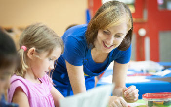 A crayola imagine arts academy instructor leaning over a girl helping her with her art project