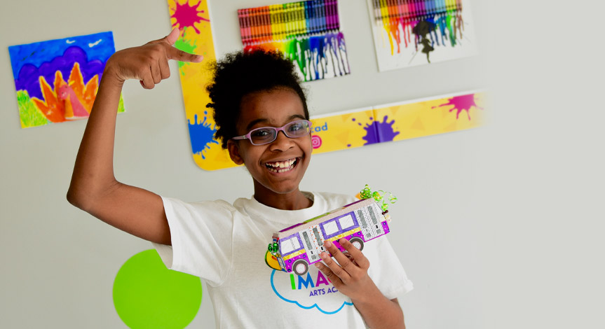 A girl with glasses is smiling while pointing at her paper bus she made during a Crayola imagine arts academy class